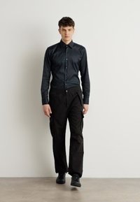 Zwarte overhemden met knopen, zwarte cargobroek met zijzakken en zwarte schoenen. Glad materiaal met een slim fit en subtiele glans.