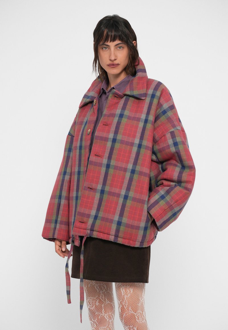 Red and blue plaid oversized jacket with a high collar, large front buttons, and a drawstring hem. Worn over a brown skirt and textured tights.