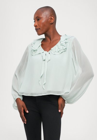 Light mint green, sheer blouse with ruffled neckline and tie detail. Features long, flowing sleeves and a cropped cut. Black jeans visible.
