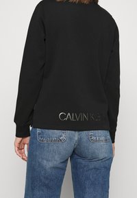 Person som bär en svart sweatshirt med "CALVIN KLEIN" tryckt i silver på ryggen, i kombination med blå jeans.