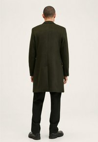 Lange, donkergroene jas van een wolmix, met een rechte snit, notch-revers en zijzakken; gecombineerd met zwarte broeken en schoenen.