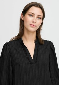 Black blouse with a V-neck, textured fabric, and puffed sleeves. Featuring intricate trim along the neckline for added detail.