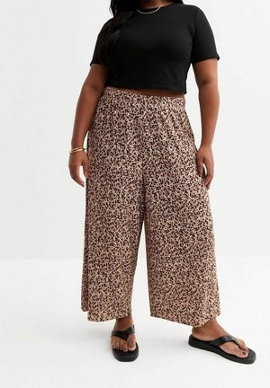 Kobieta w czarnym krótkim topie odsłaniającym brzuch, szerokich beżowych spodniach z czarnym nakrapianym wzorem, czarnych sandałach, złotej bransoletce i cienkim naszyjniku.