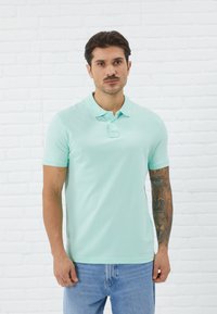 Mintgrønn poloskjorte med klassisk krage, korte ermer og to knapper. Brukt med lyseblå jeans, med arm-tatoveringer.