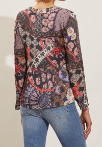 Kvinnas rygg som bär en långärmad blus med ett färgglatt abstrakt blommönster och paisleymönster i lappar, tillsammans med ljusblå jeans.