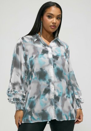 Kobieta z długimi czarnymi włosami ubrana w niebieską, szarą i białą bluzkę z efektem tie-dye, zapinaną na guziki, z marszczonymi mankietami i długimi kryształowymi kolczykami.