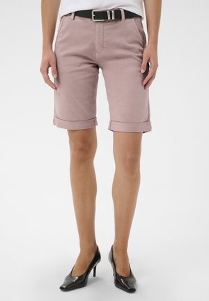 Woman wearing rolled-up mauve shorts, black belt, white top, and black high-heeled shoes, standing against a plain background.