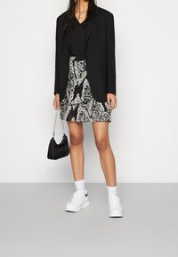 Blazer negro sobre una blusa negra de cuello en V, combinado con una falda corta con estampado. Zapatillas blancas y un bolso negro pequeño. Fondo neutro.