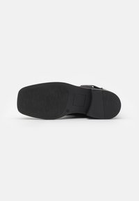 Semelle de chaussure noire présentant un motif texturé, un design plat et un léger talon. Le matériau semble durable avec un accent en métal rectangulaire.