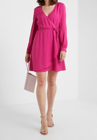 Różowa sukienka w stylu kopertowym z długimi rękawami, dekoltem w kształcie litery V i regulowanym wiązaniem. Gładki materiał o zwiewnym kroju, w zestawie z małą różową torebką.