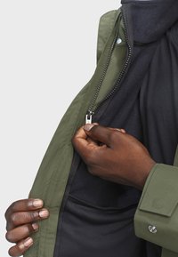 Zelená bunda se zipem vpředu, má hladký vnější povrch a měkkou fleecovou podšívku viditelnou uvnitř. Ruka držící zip.