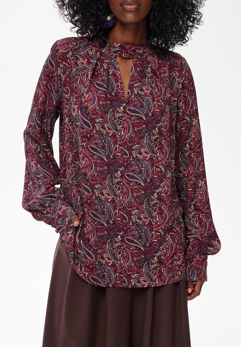 Vrouw in een bordeauxrode paisley blouse met lange mouwen en een keyhole halslijn, gedeeltelijk gestoken in een donkerbruine rok, staande tegen een witte achtergrond.