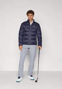 Temno modra puhovka z zadrgo spredaj, z vodoravnimi šivi, tesno prilegajoča se, nošena preko bele srajce, v kombinaciji s svetlo sivimi hlačami in golf palico.