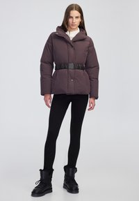 Mujer vestida con una chaqueta acolchada marrón oscuro con un cinturón negro, leggings negros y botas de invierno forradas de piel negra, de pie contra un fondo liso.