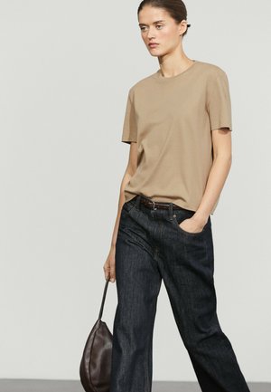 Femme portant une chemise beige à manches courtes et un jean foncé, tenant un sac en cuir marron dans une main et l’autre main dans la poche.