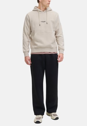 Beige hoodie with front pocket and logo, layered over a pink shirt. Paired with loose black pants and light-colored sneakers.
