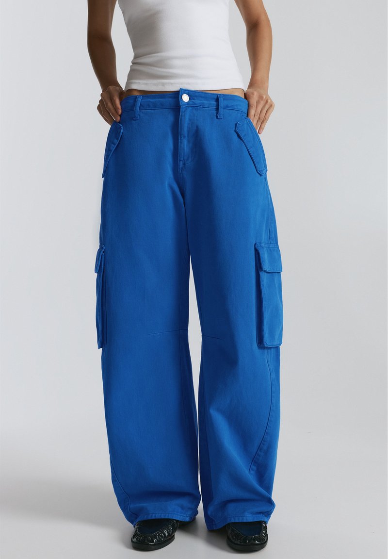 Personne portant un pantalon cargo bleu vif à jambes larges avec de grandes poches latérales et des chaussures noires, debout devant un fond clair uni.
