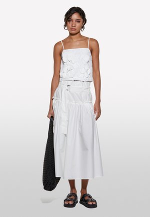 Top court blanc avec une texture florale, associé à une jupe blanche longue avec une taille élastique et un lien ajustable. Sac tissé noir.
