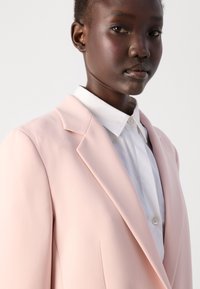 Helle rosa Blazer mit scharfen Revers, weicher Textur und strukturiertem Schnitt, getragen über einem weißen Hemd mit Kragen, das klare Linien betont.