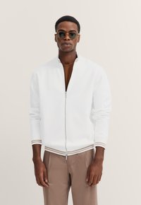 White zip-up bomber jacket with a stand collar, ribbed cuffs, and hem featuring brown and cream striped accents. Worn over a brown shirt.
