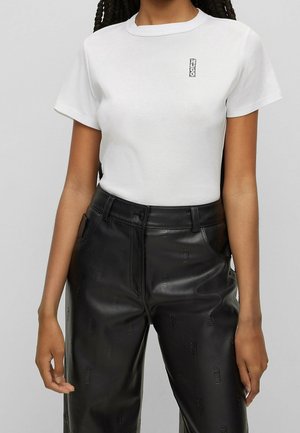 Frau trägt ein weißes, figurbetontes Kurzarmshirt mit kleinem Logo auf der Brust und schwarze, hoch taillierte Lederhosen mit eingeprägten Logos.