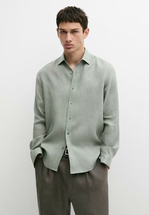 Giovane uomo con capelli corti e scuri che indossa una camicia verde chiaro con bottoni e pantaloni scuri, in piedi con le mani nelle tasche.
