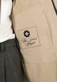 Béžová bunda s černým potiskem se švýcarským kroužkem a nápisem "The Swiss Yuppie" v kurzívě, texturovaná látka a viditelné detaily šití.