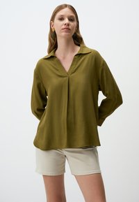 Blusa verde oliva a maniche lunghe con scollatura a V e colletto, dotata di una vestibilità comoda e una texture sottile. Indossata con pantaloni corti beige.