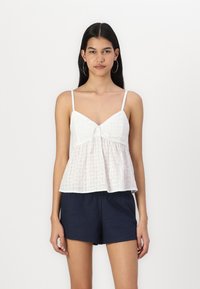 Balta, be rankovių palaidinė su dekoratyvine kaspinu ant kaklo, pagaminta iš lengvos tekstūruotos medžiagos su platėjančiu apačioje. Užsidėta su tamsiomis šortais.