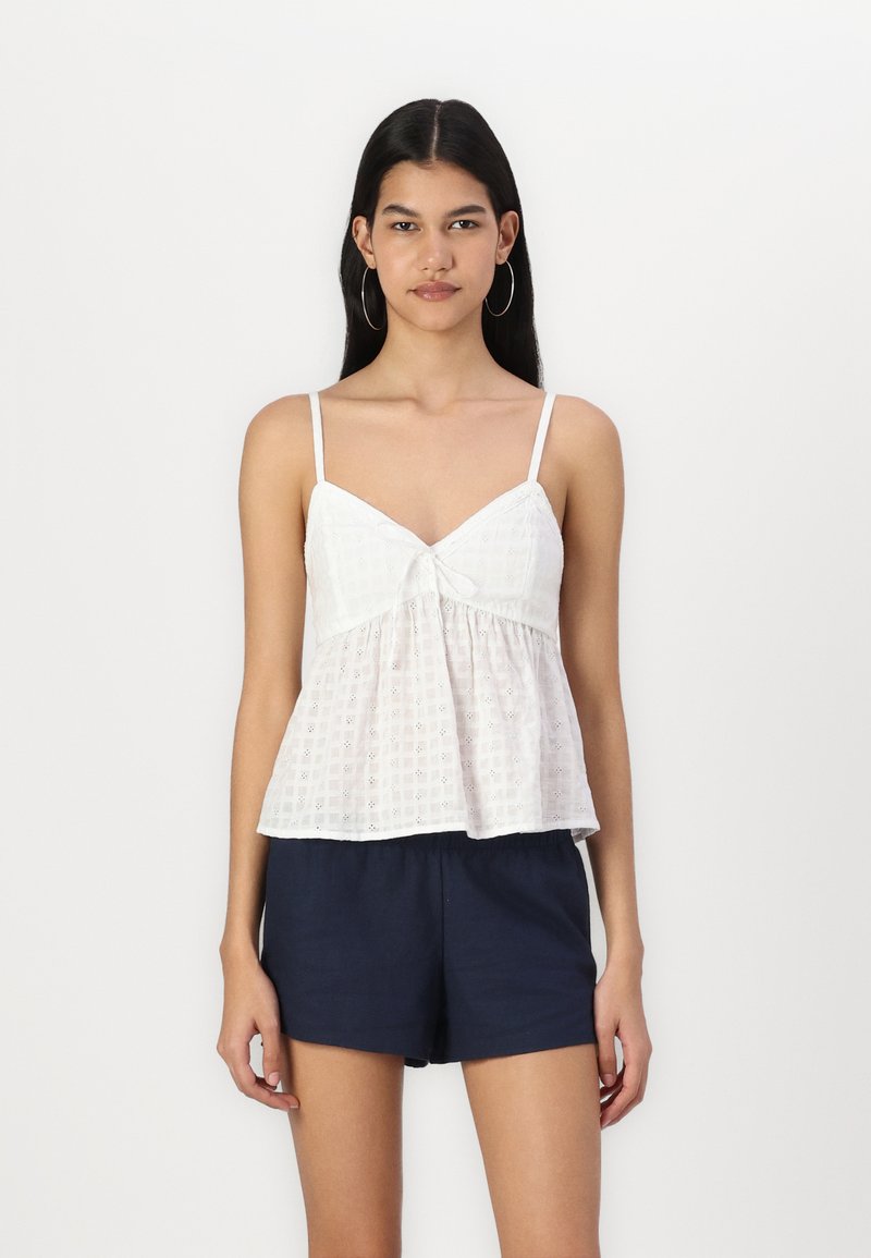 Balta, be rankovių palaidinė su dekoratyvine kaspinu ant kaklo, pagaminta iš lengvos tekstūruotos medžiagos su platėjančiu apačioje. Užsidėta su tamsiomis šortais.