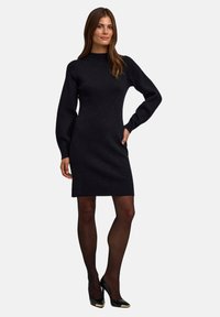 Schwarzes gestricktes Kleid mit hohem Kragen und langen Ärmeln. Hat eine figurbetonte Silhouette und wird mit schwarzen glänzenden Absätzen und Strumpfhosen kombiniert.