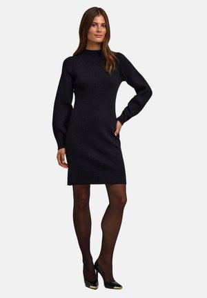 Schwarzes gestricktes Kleid mit hohem Kragen und langen Ärmeln. Hat eine figurbetonte Silhouette und wird mit schwarzen glänzenden Absätzen und Strumpfhosen kombiniert.