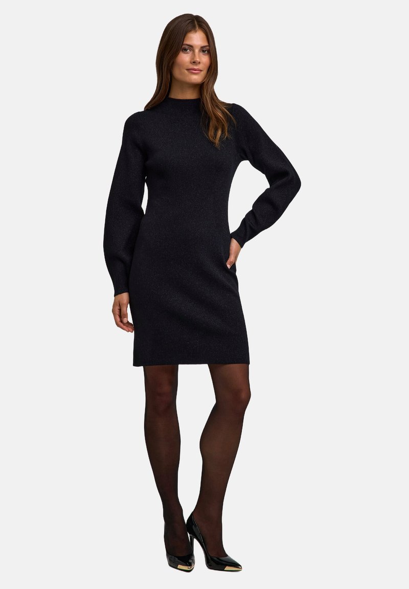 Schwarzes gestricktes Kleid mit hohem Kragen und langen Ärmeln. Hat eine figurbetonte Silhouette und wird mit schwarzen glänzenden Absätzen und Strumpfhosen kombiniert.