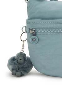 Ljusblå nylon crossbody-väska med en dragkedjeficka och en fluffig grå gorillanyckelring fäst vid den. Ytan är slät och mjuk.