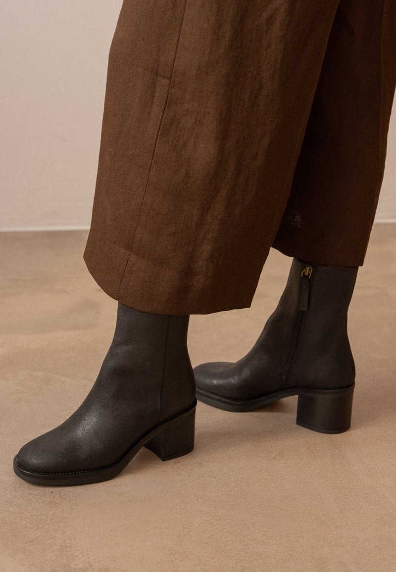 Botines de cuero negro con tacones gruesos, usados con pantalones anchos marrones recortados, sobre un suelo beige.