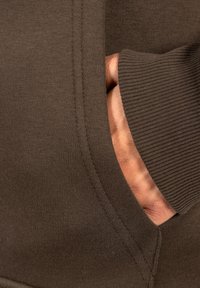 Tejido de sudadera marrón con puños acanalados y un bolsillo lateral. Una mano es visible reposando dentro del bolsillo. Acabado texturizado y suave.