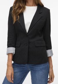 Svart skräddarsydd kavaj med slag, två framfickor och knappstängning. Ärmarna har grå och vit randig tyg. Bärs över en vit skjorta.