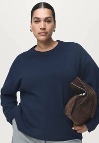 Granatowy sweter wykonany z dzianiny o luźnym kroju, okrągłym dekolcie i ściągaczach. Trzyma brązową torbę z zamszu o unikalnym, złożonym wzorze.