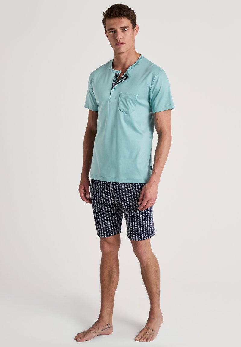 Lichtblauw overhemd met korte mouwen en een zak, met een gemusterde halslijn. Gecombineerd met donkerblauwe shorts met een subtiel herhaald patroon.