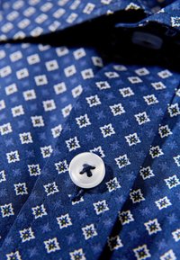 Blauw gemêleerd katoenen overhemd met een geometrisch patroon in wit en geel, met een witte knoop in het midden van de kraag. Gemaakt van gestructureerd katoen.