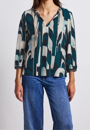 Vrouw draagt een blouse met een abstract patroon in teal, zwart en wit met driekwart mouwen en blauwe spijkerbroek tegen een effen achtergrond.