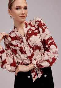 Czerwona bluzka w kwiaty z zaciąganym sznurkiem przy szyi, długimi bufkami i miękkiego materiału. Akcentowana kremowymi wzorami kwiatowymi i detalami guzików.
