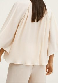 Lyse beige bluse med et flydende design og brede ærmer, der har et struktureret ribbet mønster og en subtil glans. Vist båret over matchende bukser.