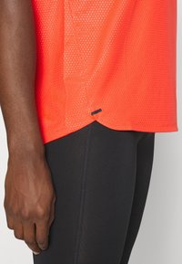 Camiseta atlética de malla roja con una superficie texturizada, con dobladillo curvado y una pequeña etiqueta negra de acento, combinada con leggings negros.