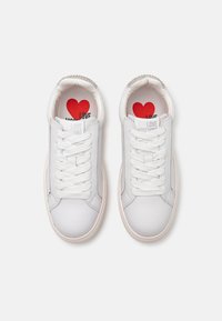 Sneakers in pelle bianca con lacci piatti, punta tonda, suola in gomma beige e grafica di cuore rosso all'interno. Presentano la scritta stampata "LOVE MOSCHINO".