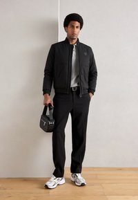 Jachetă bomber neagră cu manșete și guler tricotate, tricou polo gri, pantaloni negri, adidași albaștri și gri, ținând o geantă neagră cu model.