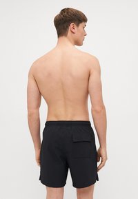 Shorts de baie negri cu un model texturat, talie elastică, buzunare laterale și un buzunar pe spate cu nasture, purtați de un model masculin văzut din spate.