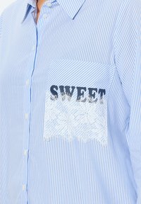 Camisa de botones a rayas azul y blanca con un bolsillo de encaje que lleva la palabra "SWEET" en letras negras. La camisa tiene un cuello clásico y una textura suave.