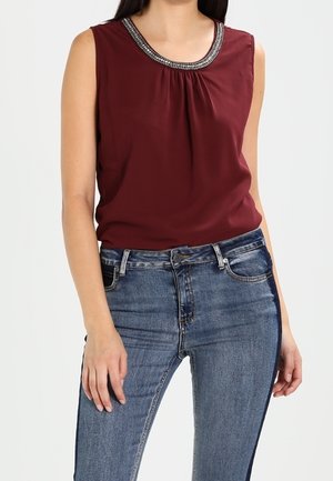 Kobieta w bordowej bluzce bez rękawów z koralikowym wykończeniem na dekolcie oraz wysokim stanem niebieskich dżinsów z bocznym paskiem.