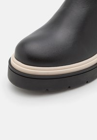 Bota negra con punta redondeada, textura de cuero liso y suela de goma beige en contraste. Detalles estriados notables en el borde de la suela.